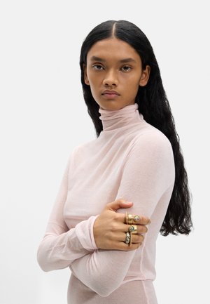 Kobieta z długimi czarnymi włosami, ubrana w jasnoróżowy sweter z golfem i nosząca na prawej ręce trzy duże złote pierścienie z kamieniami, trzyma lewą rękę.