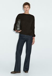 Donna con capelli corti che indossa un maglione a righe scure, pantaloni scuri e scarpe marroni, mentre tiene una borsa nera su uno sfondo chiaro.