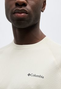 Κοντινό πλάνο ενός ατόμου που φοράει ανοιχτό μπεζ πουκάμισο Columbia, με το λογότυπο της μάρκας στο στήθος και το κάτω μισό του προσώπου ορατό.