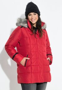 Giacca puffer rossa con cappuccio bordato in pelliccia, dettagli neri, tasche con zip e texture trapuntata. Indossata con un cappello nero e abbigliamento scuro.