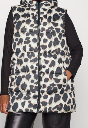 Bezruká péřová vesta s kapucí v černo-béžovém leopardím vzoru nošená přes černé tričko s dlouhým rukávem a černé kalhoty.