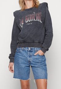 Ciemnoszara bluza z nadrukiem "LA BOHÈME", z wyściełanymi ramionami, ściągaczami na rękawach, w połączeniu z wysokim stanem krótkich jeansów. Szczegóły z postrzępionym wykończeniem.