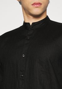 Cămașă neagră cu guler mandarin, având un material texturat și butoane negre lucioase, evidențiind un design modern și minimalist.