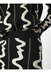 Vêtement tricoté noir avec des motifs ondulés blancs et des rayures verticales ; la taille côtelée met en valeur le design et la texture.