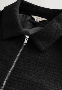 Zwarte zip-up jack van een gestructureerde stof, met een puntige kraag en zilveren rits. Binnen gevoerd met een gladde, donkere stof. M-maattag zichtbaar.