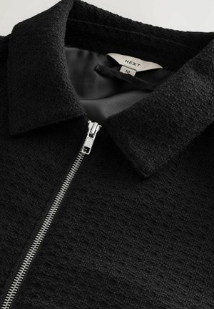 Chaqueta negra con cremallera y tejido texturizado, con cuello puntiagudo y cremallera plateada. Por dentro está forrada con un material oscuro y suave. Etiqueta de talla M visible.