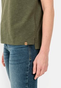 Groene T-shirt met een ronde zoom en zijslitsen, gecombineerd met blauwe denim jeans. Heeft een klein bruine logo-label aan de onderzoom.