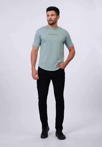 T-shirt verde chiaro a maniche corte con stampa corsiva "Los Angeles", abbinata a jeans neri slim-fit e scarpe nere. Design semplice.