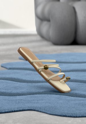 Sandalia plana beige con tres tiras delgadas, dos cuentas doradas y lazo para el dedo sobre una alfombra azul texturizada y fondo desenfocado con cojines grises.