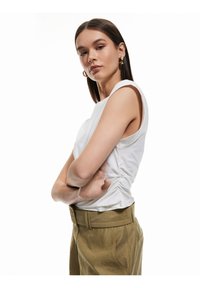 Witte mouwloze top met geplooide details aan de zijkant, gecombineerd met lichtbruine hooggetailleerde broek met een brede riem.