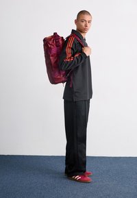Sac à dos rouge et violet avec une finition texturée, sangles réglables, porté par une personne portant un haut noir et un pantalon de survêtement, se tenant sur un tapis bleu.