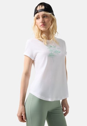 Junge Frau mit schwarzer Cap, die rückwärts getragen wird, weißem T-Shirt mit grünem Motiv und hellgrünen Leggings, die seitlich schaut vor einem einfarbigen Hintergrund.