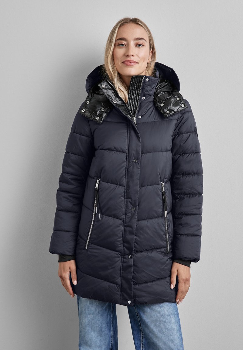 Abrigo tipo puff combinado en navy con una capucha texturada de patrón negro. Presenta bolsillos laterales con cremallera y cierres a presión. Tejido suave y acolchado.