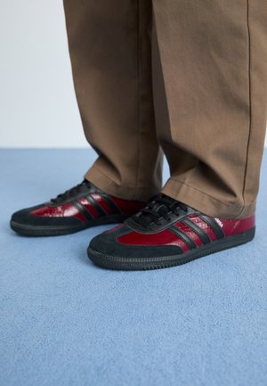 Osoba nosząca brązowe spodnie i czarno-ciemnoczerwone trampki stojąca na jasno niebieskim dywanowym podłożu.