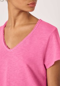 Približnji posnetek vratu in ramena ženske, ki nosi roza T-shirt z V-izrezom, obrobe izreza in rokavov so neravno zaključene.