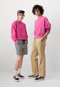 Deux personnes se tiennent côte à côte, portant des sweat-shirts rose vif avec des chaussures à carreaux noirs et blancs ; l'une porte un short à carreaux et une casquette, l'autre un pantalon beige et des lunettes de soleil.
