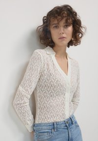 Blusa in pizzo a maniche lunghe color avorio con chiusura a bottoni e colletto a punte, caratterizzata da intricati motivi floreali e tessuto testurizzato.