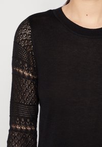 Pull noir avec un corps uni et des détails en dentelle complexes sur la manche gauche, doté d'un col rond et d'un matériau en maille texturée.