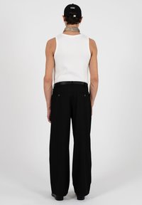 Homme de dos portant un débardeur blanc, un pantalon large noir, une casquette noire, un foulard gris autour du cou et une ceinture noire sur un fond uni.