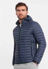 Giacca puffer blu navy dal design aderente, texture trapuntata, zip principale e cappuccio integrato. Presenta tasche laterali con zip e dettaglio del logo.