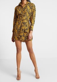 Frau, die ein gelb-schwarz gestreiftes Tigerhemdkleid mit langen Ärmeln und klaren, ankle-strap Absätzen trägt, steht vor einem weißen Hintergrund.