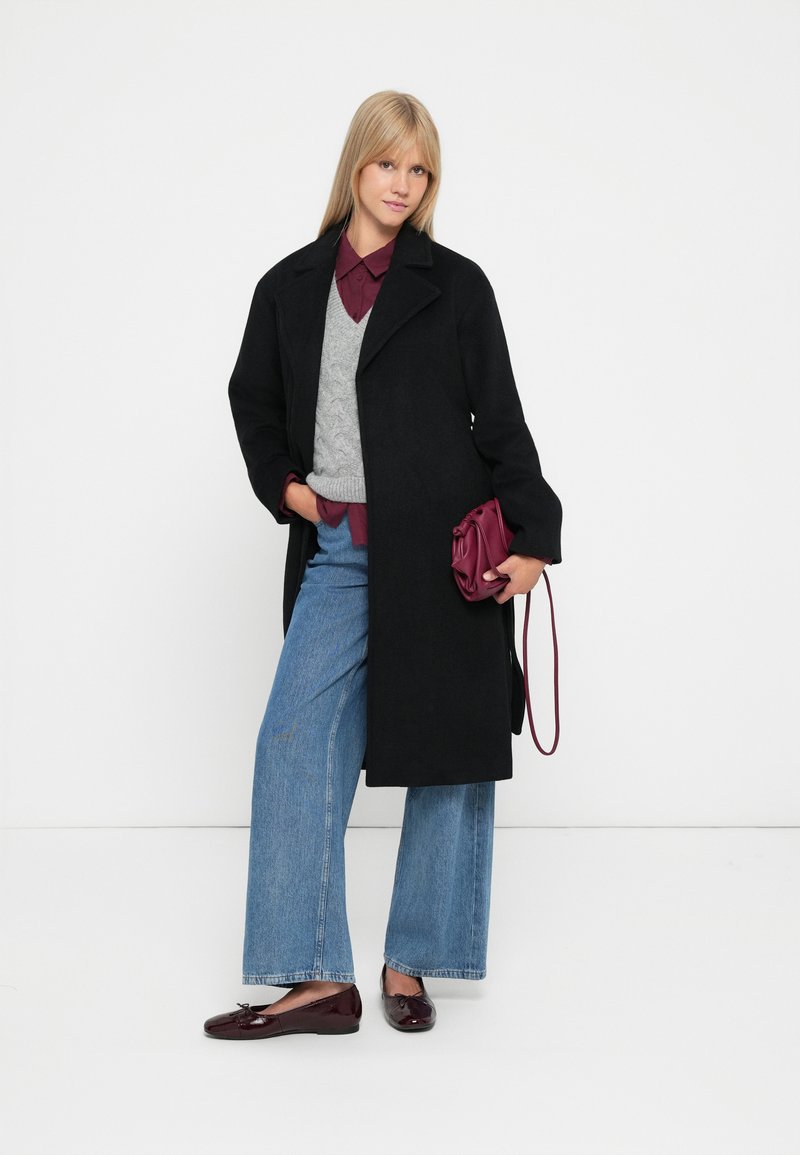 Svart ullrock med öppen front och breda slag, lager-på-lager över en grå kabelstickad tröja och bordeauxskjorta, kombinerad med vida jeans och bordeauxväska.