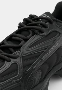 Black athletic shoe featuring a textured upper with rubber accents, rounded toe, and lace-up design. "LACOSTE" branding visible.