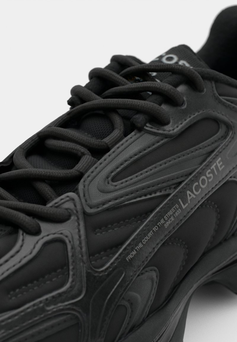 Black athletic shoe featuring a textured upper with rubber accents, rounded toe, and lace-up design. "LACOSTE" branding visible.