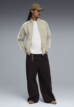 Mand med langt hår iført beige kasket, beige Puma træningsjakke, hvid skjorte, sorte bukser med vide ben og nøgler fastgjort, samt brune sko, stående med hænderne i lommerne.