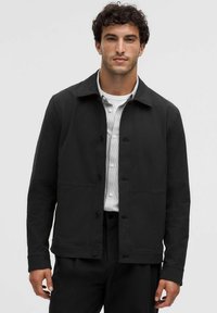 Hombre vestido con una chaqueta negra abotonada sobre una camisa blanca con rayas, de pie frente a un fondo claro y liso.