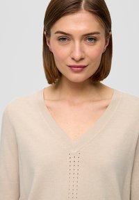 Beiger V-Ausschnitt-Pullover mit geripptem Ausschnitt, der vertikale Nahtdetails entlang der Mitte aufweist. Weiche Textur und entspannte Passform.