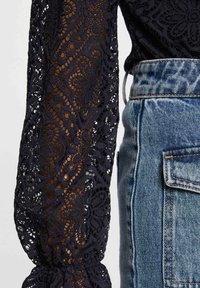 Blusa di pizzo blu navy con motivi floreali, maniche lunghe trasparenti arricciate ai polsini, abbinata a una gonna di jeans a vita alta con tasca posteriore.