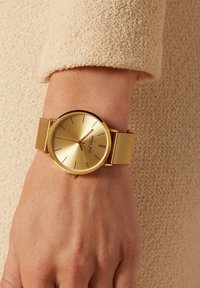 Gold watch with a mesh band, minimalist design, round face, and slender hour markers. Smooth texture; reflective surface.