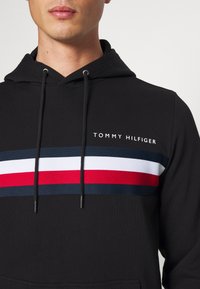 Czarna bluza z kapturem z przednim wzorem w pasy w kolorze granatowym, białym i czerwonym, z regulowanym sznurkiem ściągającym oraz napisem logo "TOMMY HILFIGER" nad pasem.