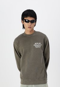 Sudadera verde oliva hecha de tejido suave, con cuello redondo de canalé y un logo gráfico blanco en el pecho. Llevada con gafas de sol negras.