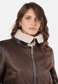Chaqueta de cuero marrón con cuello de borrego, con un cierre dorado y dos bolsillos frontales. Textura suave con color de cuello en contraste.