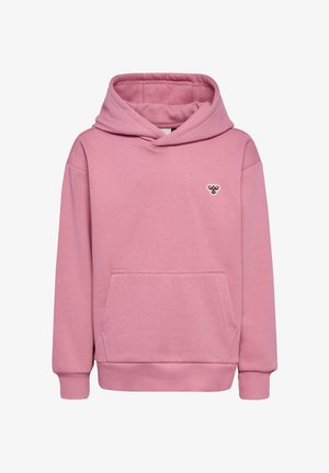 Jong meisje met blond haar, gekleed in een roze hoodie en joggingbroek, staand met handen in de zakken en witte sneakers op een effen achtergrond.