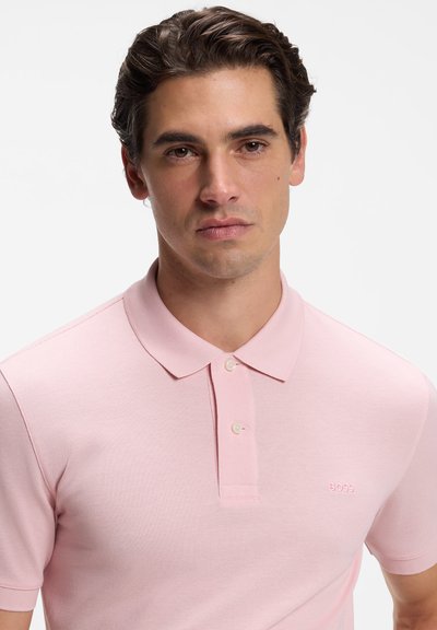 Homme aux cheveux bruns ondulés portant un polo rose clair à manches courtes avec deux boutons et un petit logo "BOSS" sur la poitrine.