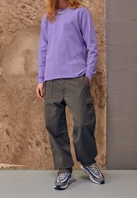 Lila långärmad skjorta, grå baggy cargo-byxor och grå sneakers med blå accenter. Texturerad bakgrund av betongvägg.