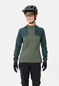 Grünes longsleeve Technikshirt mit Kapuze, kontrastierendem Brustpanel und glattem Stoff. Kombiniert mit schwarzen Handschuhen und Sporthosen.