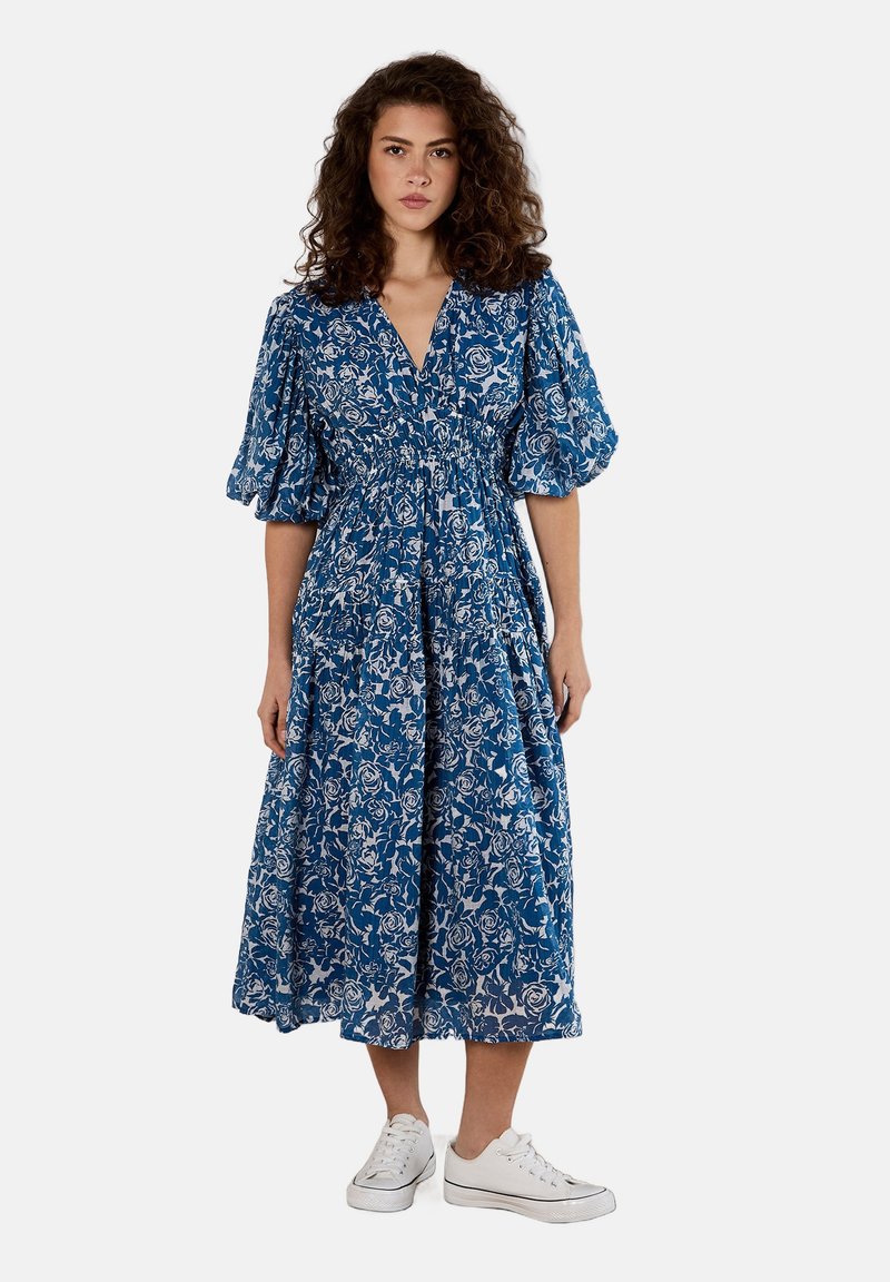 Blaues Blumenkleid mit V-Ausschnitt, puffartigen Ärmeln und elastischem Bund. Weiße Rosenmuster auf blauem Hintergrund. Kombiniert mit weißen Sneakers.