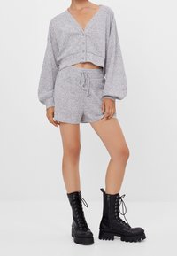 Woman wearing light gray knit cropped cardigan and matching shorts paired with black lace-up combat boots against a plain white background.