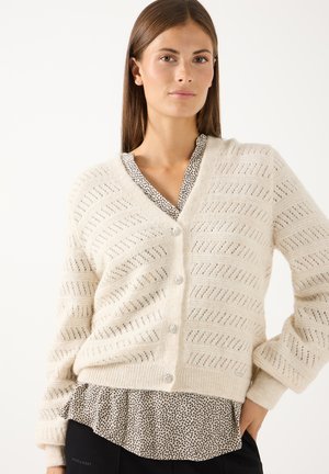 Frau trägt eine cremefarbene, strukturierte, knöpfbare Strickjacke über einer gemusterten Bluse und schwarzen Hosen, steht mit einer Hand auf der Hüfte.