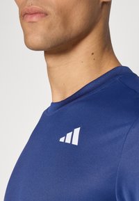 Granatowa koszulka sportowa o gładkiej fakturze, z okrągłym dekoltem i białym logo z trzema wznoszącymi się liniami na lewej stronie klatki piersiowej.