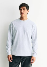 Světlé šedé tričko s dlouhým rukávem vyrobené z bavlny, s kulatým výstřihem, uvolněným střihem a žebrovanými manžetami, v kombinaci s tmavými kalhotami.