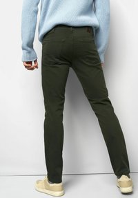 Person, der eine dunkelgrüne Slim-Fit-Hose, einen hellblauen Strickpullover und beigen Turnschuhe trägt, steht vor einem weißen Hintergrund.