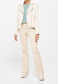 Jasno-beżowa kurtka motocyklowa z teksturowanym wykończeniem, noszona na turkusowym swetrze i dopasowanych kremowych spodniach, dopełniona neutralnymi trampkami.
