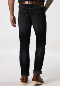 Jeans in denim blu scuro con vestibilità slim, caratterizzati da cuciture a contrasto e risvolti. Stivali in pelle marrone indossati, con decorazioni minime.