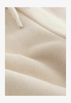 Lichtbeige katoenen stof met een zachte textuur. Het materiaal is gebreid met een lichte rek, met subtiele variaties in kleur en weefpatroon.