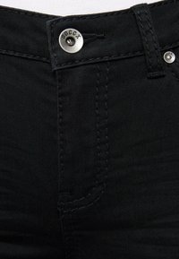 Jeans de mezclilla negra con cierre de botón de metal, costuras en contraste y una textura suave. Vista en primer plano de la zona de la cinturilla.